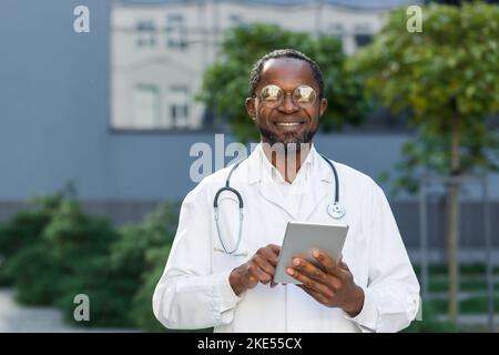 Porträt eines glücklichen afroamerikanischen Arztes mit Tablet-Computer, eines älteren Mannes, der lächelt und vor der modernen Klinik mit Brille und medizinischem Mantel auf die Kamera schaut. Stockfoto
