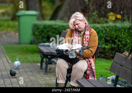 Riga, Lettland - 4. November 2022: Weibchen auf einer Bank, die Tauben aus den Händen füttert Stockfoto