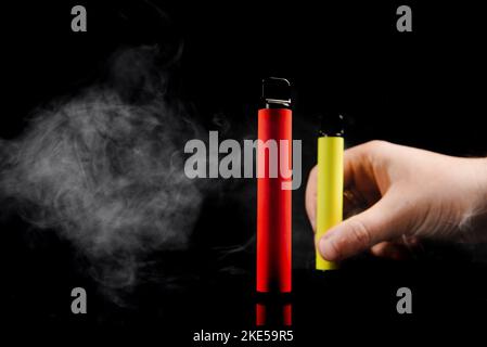 Elektronische Einweg-Zigaretten verschiedener Geschmacksrichtungen in der Hand auf einem gelben Hintergrund. Das Konzept von modernem Rauchen, Verdampfen und Nikotin. Stockfoto