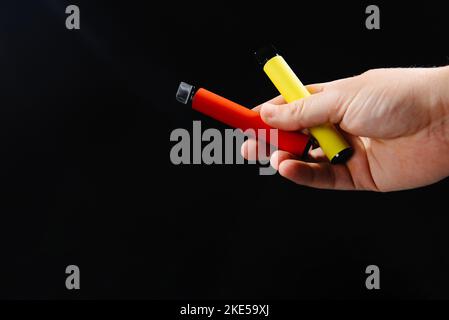 Elektronische Zigaretten in der Hand auf schwarzem Hintergrund mit leerem Raum. Elektronische Einweg-Zigaretten verschiedener Geschmacksrichtungen in der Hand auf einem gelben Stockfoto