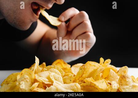 Der Mann isst Junk Food, Chips auf dunklem Hintergrund. Schädliche Lebensmittel. Frittierte Lebensmittel sind schlecht für Ihre Gesundheit. Gesunde Lebensweise, richtige Ernährung, Verbot Stockfoto