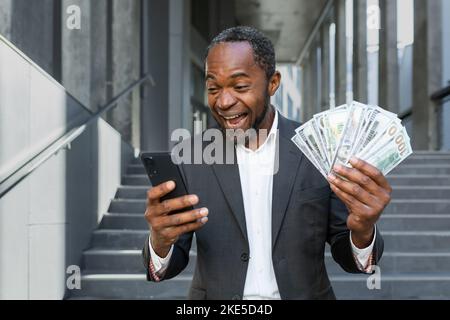 Glücklicher afroamerikanischer Geschäftsmann vor dem Bürogebäude, der Online-Gewinn und -Gewinn feiert, Mann im Geschäftsanzug, der Smartphone und Bargelddollar hält. Stockfoto