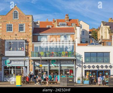 Ein typischer Blick auf ein Fish and Chip Restaurant am Meer. Die Leute sitzen auf einer Bank und essen und ein blauer Himmel ist oben. Stockfoto