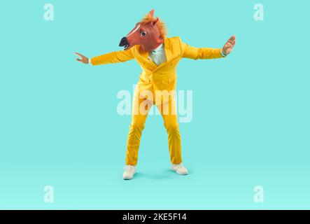 Lustiger glücklicher Mann in gelbem Anzug und Pferdemaske tanzt auf türkisfarbenem Studiohintergrund Stockfoto
