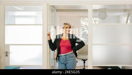 Glückliche junge Geschäftsfrau, die fröhlich lächelt, während sie vor ihrem Büro steht. Erfolgreiche Geschäftsfrau, die an einem kreativen Arbeitsplatz arbeitet. Stockfoto