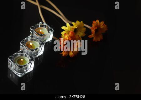 Nahaufnahme von drei stilvollen Glaskerzen in diagonaler Reihe und drei bunten Gerbera-Blüten auf schwarzer Oberfläche Stockfoto