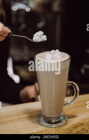 Selektiver Fokus der weiblichen Hand halten Löffel mit weißen Marshallows in der Nähe Tasse Kaffee Latte auf Holztisch auf Hintergrundbeleuchtung Girlande in modernen Coff Stockfoto