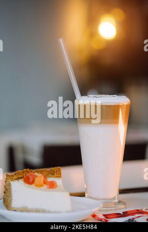 Nahaufnahme von Kuchen und großer Tasse Milchshake mit Stroh auf weißem Tisch vor Hintergrund Girlanden im Café. Konzept festliche Getränke und glückliches neues Jahr Stockfoto