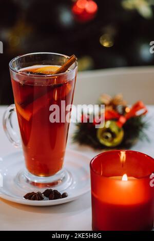 Traditionelles warmes Getränk zur Weihnachtszeit. Urlaubskonzept. Nahaufnahme von Kerze und Glühwein mit aromatischen Gewürzen und Zitrusfrüchten auf weißem Tisch Stockfoto