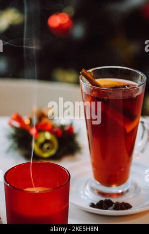 Vertikale Bilder von Kerze und weihnachten Glühwein mit Gewürzen und Früchten am Tisch auf Hintergrund geschmückt Weihnachtsbaum. Traditionelles warmes Getränk. Happ Stockfoto