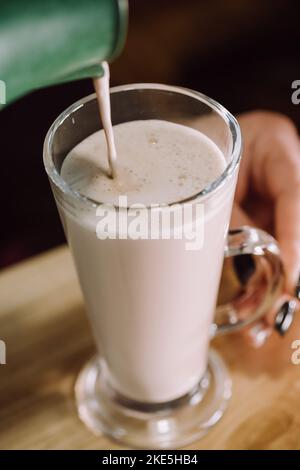 Nahaufnahme des Barista gießt geschlagene Milch in eine Tasse Kaffee. Die Kellnerin im Café serviert dem Gast an der Theke im Café einen vereisten Matcha Latte. Klein Stockfoto