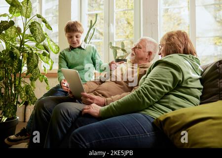 Glückliche, freundliche Familie, Großeltern und Enkel sitzen auf dem Sofa und verbringen Zeit miteinander, mit modernen Geräten, reden, studieren Stockfoto