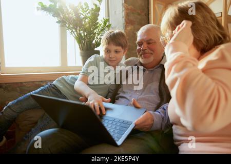 Alte Großeltern mit Enkelkind sitzen zu Hause auf der Couch und lesen Nachrichten, lustige Geschichten. Rollen auf dem Laptop ansehen. Schöne Zeit in freundlich und gemütlich Stockfoto