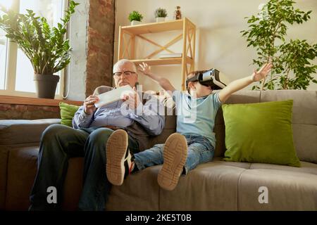 Porträt eines glücklichen älteren Mannes und kleinen Jungen, Großvaters und Enkels mit einer VR-Brille, die zu Hause auf dem Sofa sitzt und lacht. Hobbys, Freizeit Stockfoto