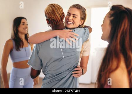 Zwei glückliche Freunde begrüßen sich gegenseitig mit einer Umarmung in einem Yoga-Studio. Gruppe von Personen, die einen Fitnesskurs in einem Gemeinschaftsstudio besuchen. Diverse p Stockfoto