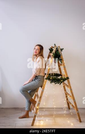 Portrait einer modernen Frau mittleren Alters an einer Holzleiter, geschmückt mit Tannenzweigen, Girlande für Weihnachten. Platz für Text, helle Wand. Skandinavische St. Stockfoto