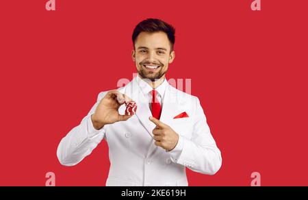 Schöner glücklicher Mann hält Poker-Chip in einer Hand und zeigt auf sie mit dem Finger der anderen Hand stehen auf rotem Studio-Hintergrund, Copyspace . Kasino Stockfoto