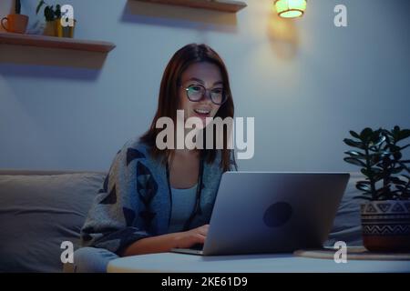 Junge lächelnde Frau, die nachts oder abends einen Laptop benutzt, Videoanruf, der zu Hause auf dem Sofa sitzt Stockfoto