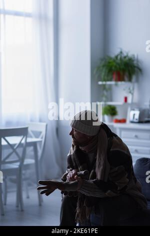 Gefrorener Mann in Strickmütze, der warme Handschuhe trägt, während er unter der Decke sitzt und in der Dämmerung wegschaut Stockfoto