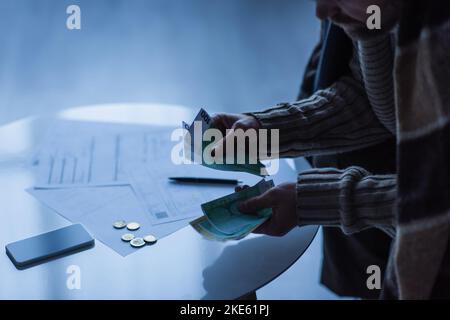 Teilansicht des Mannes, der Geld in der Nähe des Smartphones zählt, und der Zahlungsrechnungen in der Dämmerung Stockfoto