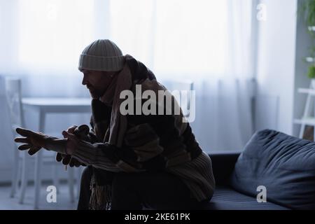 Seitenansicht eines gefrorenen Mannes, der Handschuhe anziehen musste, während er auf der Couch in einem Hut und einer warmen Decke saß Stockfoto