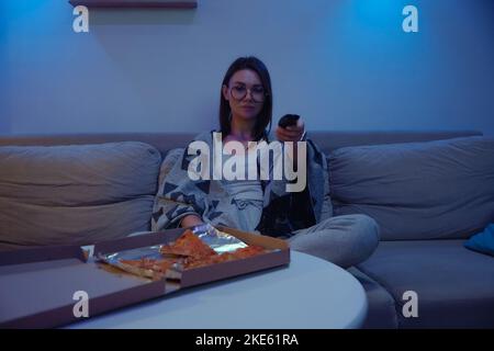 Glückliche Frau, die abends Pizza isst und fernsieht, während sie zu Hause auf dem Sofa sitzt Stockfoto
