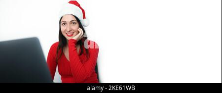 Schöne lächelnde Brünette Frau in Silvester Weihnachtsmann Hut funktioniert mit Laptop. Bürokonzept Weihnachten und Neujahr. Junge Freiberuflerin, ganz rot in fröhlicher Stimmung gekleidet Stockfoto