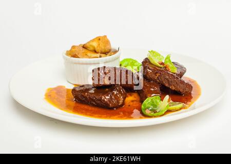 Gourmet Kalbsbacken in roter Sauce mit Grün. Nahaufnahme auf einer weißen Platte auf weißem Hintergrund Stockfoto