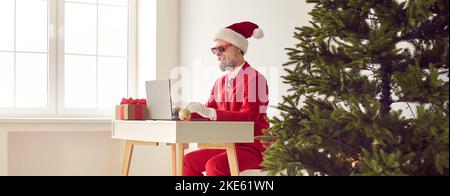 Fröhlicher grauhaariger bärtiger Mann in rotem Anzug, Weihnachtsmann-Mütze sitzt am Schreibtisch und arbeitet auf einem Laptop in der Nähe des Weihnachtsbaums. Stockfoto