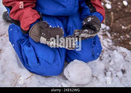 Knabe, der im Gebäude einen Schneeball kniet, bekleidet mit einem Schneeanzug und Handschuhen. Stockfoto