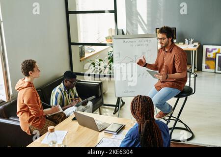 Junger, selbstbewusster Business Coach, der mit Grafiken am Whiteboard sitzt und der Gruppe interkultureller Kollegen beim Seminar die Daten erklärt Stockfoto