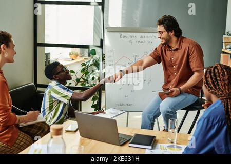 Zwei junge, erfolgreiche interkulturelle Geschäftsleute, die nach der Präsentation beim Seminar mit Diagrammen gegen das Whiteboard schlagen Stockfoto