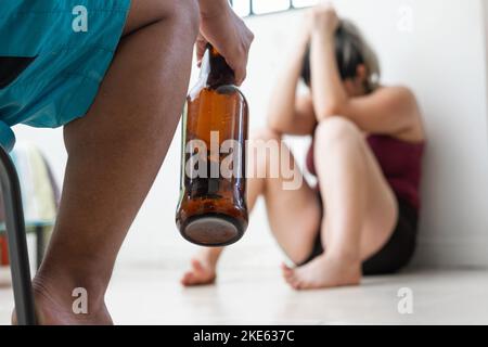 Nahaufnahme eines Mannes, der eine Flasche Bier in der Hand hält, im Hintergrund seine Frau, die weinend in fetaler Position auf dem Boden sitzt Stockfoto