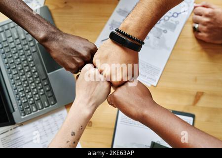 Über dem Winkel von vier Händen junger interkultureller Mitarbeiter, die sich mit Laptop und Finanzdokumenten über den Tisch schlagen Stockfoto