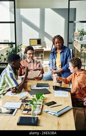Eine Gruppe junger Mitarbeiter, die am Tisch im Büro versammelt waren, um Kaffee zu trinken und einige Arbeitspunkte bezüglich des neuen Geschäftprojekts zu besprechen Stockfoto
