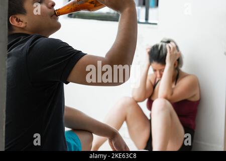 lateinischer Mann trinkt ein Bier, während seine Frau weinend auf dem Boden sitzt. Häusliche Gewalt in einem jungen Haus Stockfoto