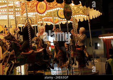 Ein traditionelles Karussell auf dem Messegelände wird nachts beleuchtet, während Erwachsene Männer und Frauen eine Fahrt auf dem Karussell in York, North Yorkshire, England, Großbritannien genießen. Stockfoto