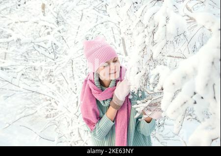 Junge Frau auf dem Hintergrund einer schönen Pflanze im Schnee. Eine junge Dame berührt bei einem Schneefall einen Ast im Schnee. Tolle Stimmung, Freude, Spaß. Cop Stockfoto
