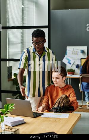 Ein junger, selbstbewusster Geschäftsmann in einem gestreiften Hemd erklärt dem Kollegen die Daten, während er während der Diskussion auf den Laptop-Bildschirm zeigt Stockfoto