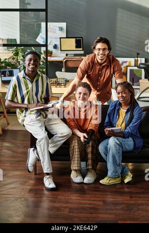 Team von jungen, fröhlichen multikulturellen Managern, die im Büro mit Computern und anderen Vorräten auf der Couch sitzen und an ihren Arbeitsplätzen sitzen Stockfoto