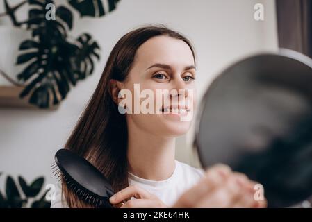 Fröhliche, attraktive junge Dame sitzt zu Hause auf der Couch, kämmt ihr schönes seidiges Haar mit einem Holzpinsel, schaut auf den Spiegel und lächelt. Niedliche Hündin tun Stockfoto