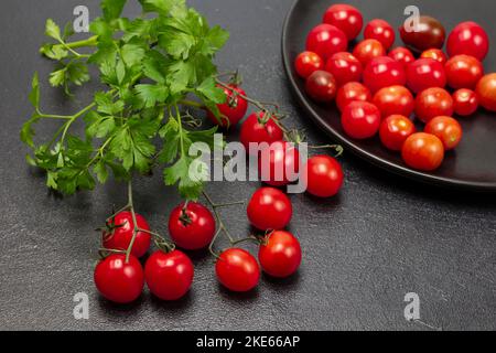 Petersilienzweig und Kirschtomaten auf dem Tisch. Kleine rote Tomaten auf schwarzem Teller. Draufsicht. Schwarzer Hintergrund Stockfoto