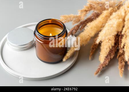 Brennende Duftkerze in einem dunklen Glasgefäß mit dem Aroma von Gewürzen. Herbststimmung mit trockenem Schilfgras und Betonplatte. Gemütlicher Lebensstil Stockfoto