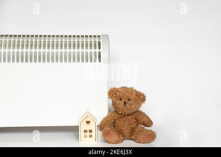 Elektrischer Konvektor, ein kleines Holzhaus und ein Teddybär daneben auf weißem Hintergrund, ein warmes Zuhause Stockfoto