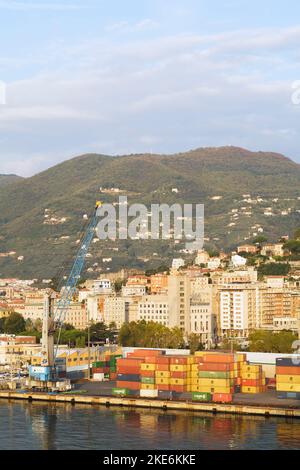 Gestapelte Transportcontainer und blauer Portalkran auf dem Dock im Handelshafen von La Spezia und der Skyline der Stadt, Italien Stockfoto