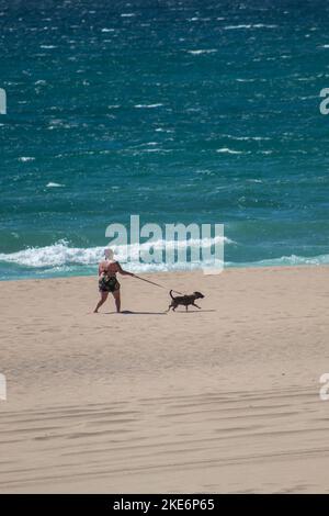 Eine übergewichtige Frau im Bikini, die ihren Hund an der Leine am Sandstrand Playa del Guincho in Spanien führt Stockfoto