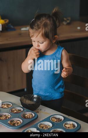 Nettes kleines glückliches Baby Mädchen, das Muffins mit Blaubeeren in der heimischen Küche dekoriert, drinnen. Lustige schöne gesunde Kind Spaß, Geschmack süß Stockfoto