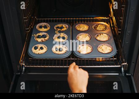 Frau Hände nehmen aus dem heißen Ofen leckere Blaubeeren Cupcakes in der Küche. Junge Erwachsene Mädchen kochen Gebäck zu Hause. Menschen leben. Süßes Essen. Still Stockfoto