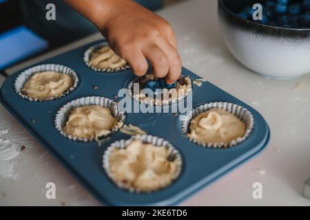 Nahaufnahme der Kinderhände, die die noch rohen Muffins mit Blaubeeren dekorieren. Lustige schöne gesunde Kind Spaß mit helfen. Eltern helfen Stockfoto