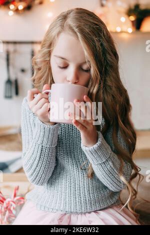 Ein blondes Mädchen mit langen Haaren trinkt mit Vergnügen Kakao aus einer rosa Tasse. Weihnachtliche Atmosphäre. Stockfoto